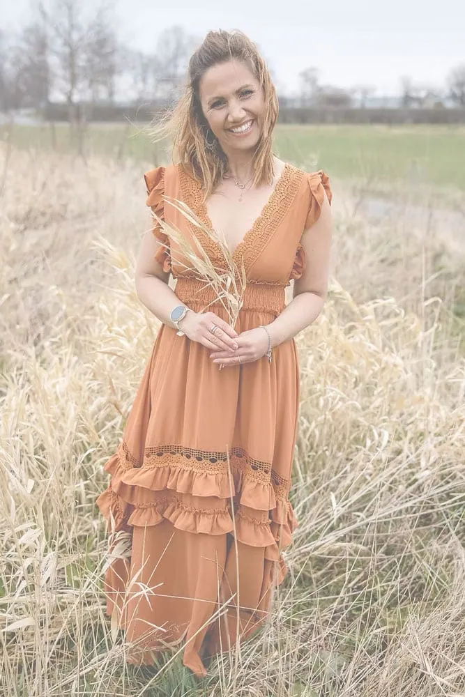 Eine lächelnde Frau steht auf einem Feld mit trockenem Gras und hält einige Grashalme in ihren Händen.